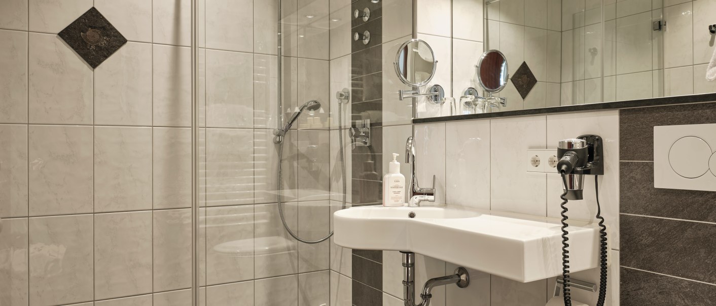 Modernes Badezimmer mit großer Dusche, Waschbecken, Spiegel und Toilette. Helle Fliesen und dunkler Boden sorgen für einen eleganten Look., © Badhotel Restaurant Stauferland