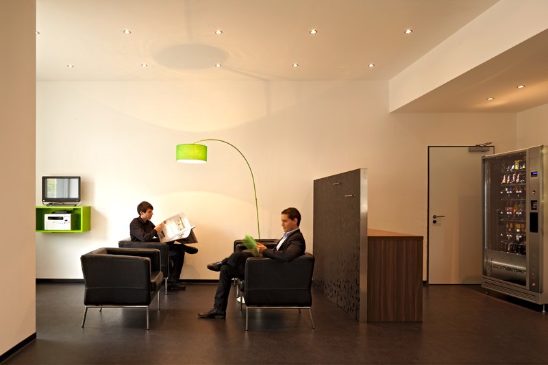 Modern lobby with two men in black armchairs, reading material in hand, green floor lamp, vending machine in the background., &copy; a2 HOTELS