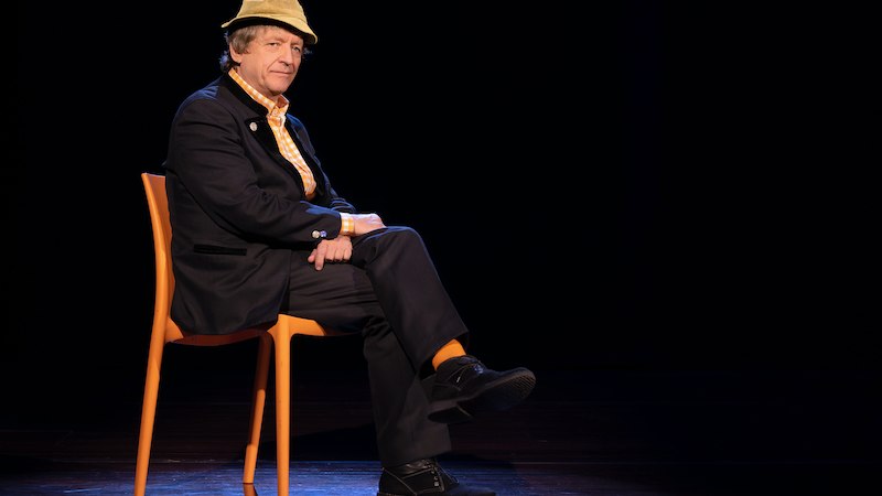 Eine Person mit Hut sitzt auf einem orangefarbenen Stuhl auf einer dunklen Bühne. Sie trägt einen Anzug und schaut in die Kamera., © Theaterhaus Stuttgart e.V.