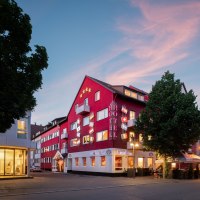 Rotes Hotelgeb&auml;ude mit Flaggen und beleuchteten Schildern bei D&auml;mmerung, umgeben von B&auml;umen und Stra&szlig;enlaternen., &copy; Hetzel Hotel Stuttgart