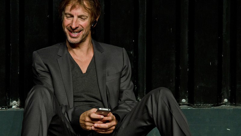 Ein Mann in einem Anzug sitzt lächelnd mit einem Smartphone in der Hand vor einem dunklen Hintergrund., © Theaterhaus Stuttgart e.V.