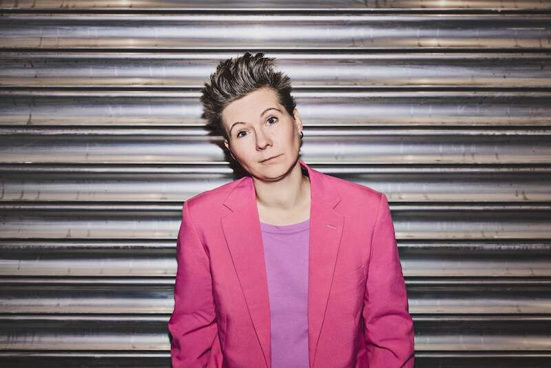 A person with short hair is wearing a pink blazer and a purple shirt. She stands in front of a metallic wall and looks directly into the camera., &copy; Rosenau Kultur e.V.