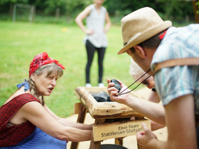 Eine Frau und ein Mann bei einer Outdoor-Aktivität. Der Mann hält eine Stoppuhr, die Frau arbeitet an einer Holzkonstruktion. Im Hintergrund eine weitere Person., © Cool-Tours StattReisen Eine Frau und ein Mann bei einer Outdoor-Aktivität. Der Mann hält eine Stoppuhr, die Frau arbeitet an einer Holzkonstruktion. Im Hintergrund eine weitere Person., © Cool-Tours StattReisen