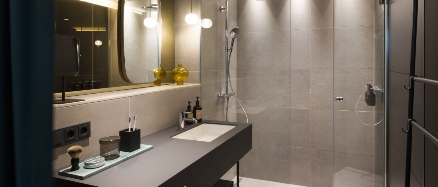 Modernes Badezimmer mit großer Dusche, stilvollem Waschbecken und beleuchtetem Spiegel. Dezente Beleuchtung und elegante Einrichtung., © EmiLu Hotel GmbH