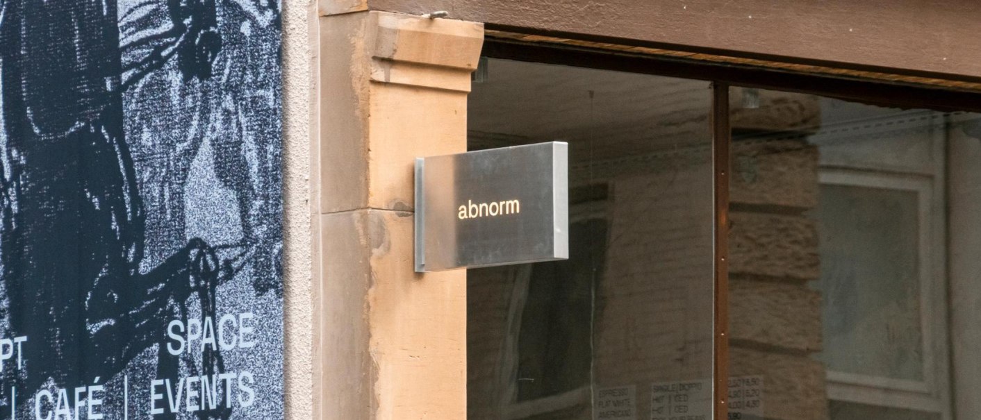 Ein Metallschild mit der Aufschrift 'abnorm' h&auml;ngt an einer Wand. Daneben ist ein Kunstplakat zu sehen., &copy; SMG, Sarah Schmid