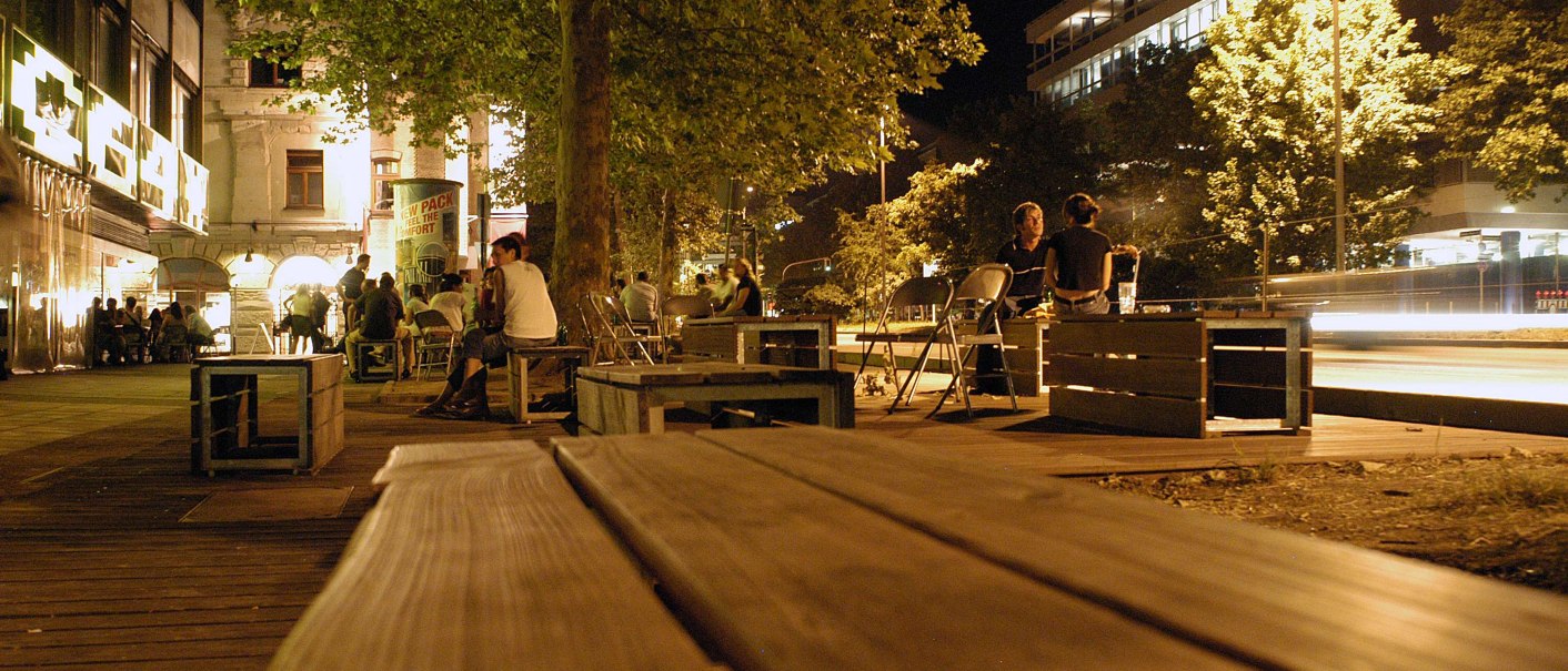 Nachtaufnahme der Theodor-Heuss-Straße in Stuttgart. Menschen sitzen auf Holzbänken unter Bäumen, umgeben von beleuchteten Gebäuden und Straßenlaternen., © Stuttgart-Marketing GmbH, Marc Gilardone Nachtaufnahme der Theodor-Heuss-Straße in Stuttgart. Menschen sitzen auf Holzbänken unter Bäumen, umgeben von beleuchteten Gebäuden und Straßenlaternen., © Stuttgart-Marketing GmbH, Marc Gilardone