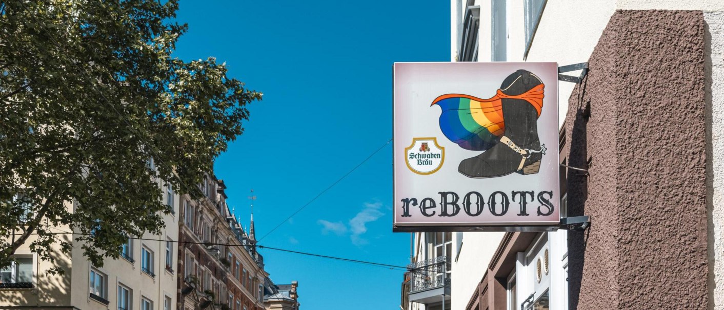 Ein Schild mit einem Stiefel und einer Regenbogenflagge, daneben die Aufschrift 'reBOOTS'. Im Hintergrund sind Gebäude und ein Baum zu sehen., © SMG, Sarah Schmid