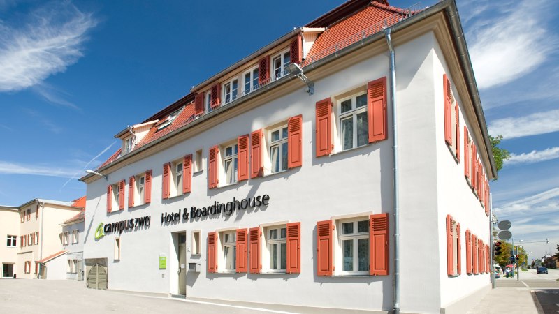 Wei&szlig;es Geb&auml;ude mit roten Fensterl&auml;den, blauer Himmel. Schriftzug: 'campuszwei Hotel & Boardinghouse'. Moderne Architektur, sonniger Tag., &copy; TOMAS