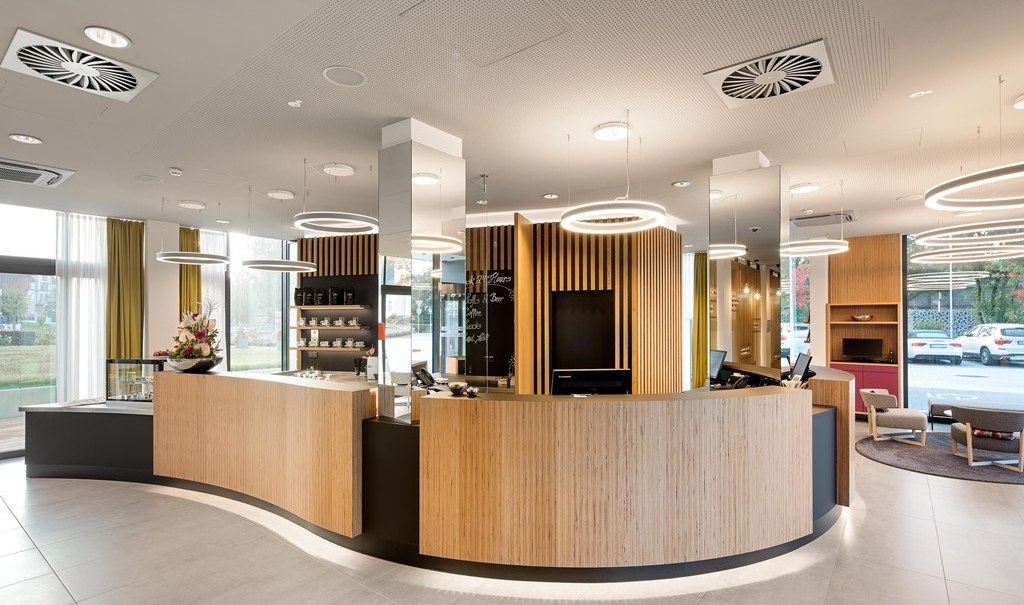 Modern reception desk with wooden paneling, round ceiling lights and glass front. Shelves with cups and a flower arrangement can be seen in the background., © Hotel am Remspark Modern reception desk with wooden paneling, round ceiling lights and glass front. Shelves with cups and a flower arrangement can be seen in the background., © Hotel am Remspark