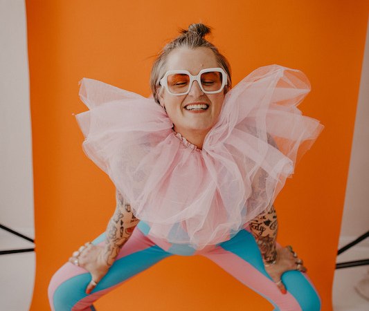 Eine Person in bunter Kleidung und großem Tüllkragen posiert fröhlich vor einem orangefarbenen Hintergrund., © Rosenau Kultur e.V. Eine Person in bunter Kleidung und großem Tüllkragen posiert fröhlich vor einem orangefarbenen Hintergrund., © Rosenau Kultur e.V.