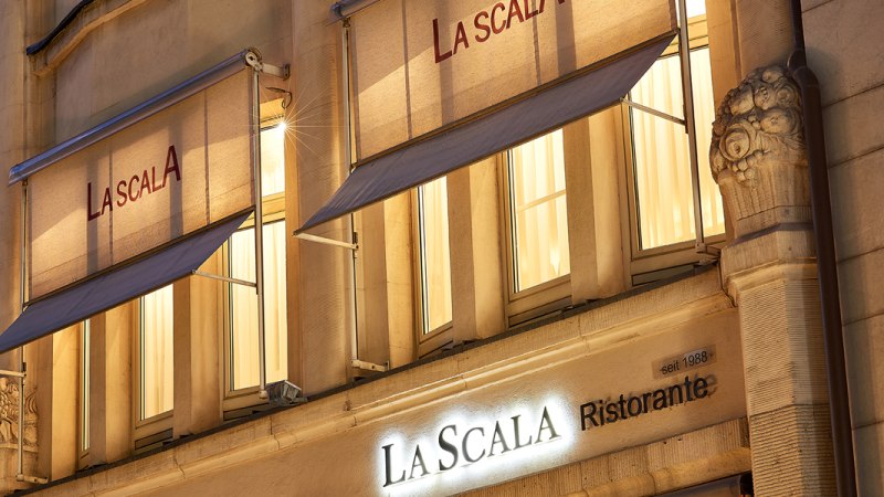Illuminated façade of the La Scala restaurant in Stuttgart with awnings and decorative columns at night., © La Scala