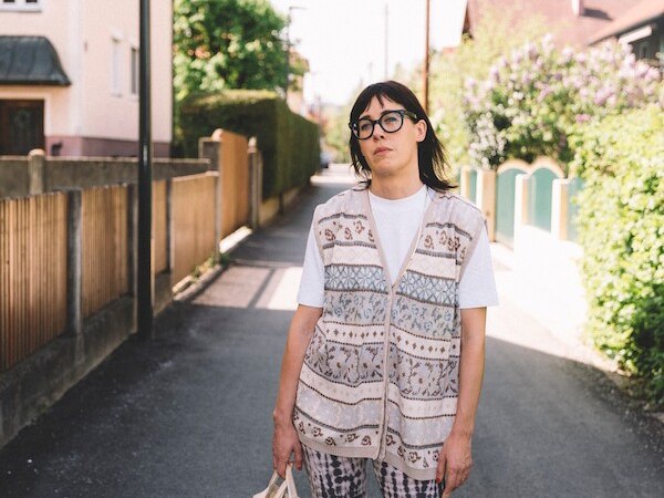 Person with glasses and patterned vest stands on a quiet street with a shopping bag. Houses and bushes can be seen in the background., &copy; Rosenau Kultur e.V.