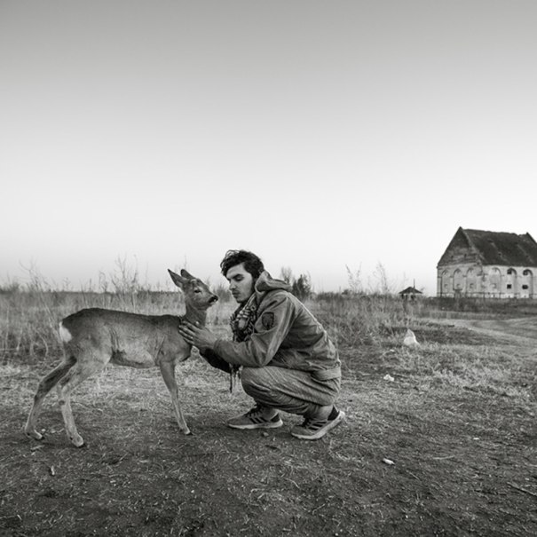 A man kneels in a field and strokes a deer. An old building can be seen in the background. The scene appears calm and peaceful., &copy; Theaterhaus Stuttgart e.V.