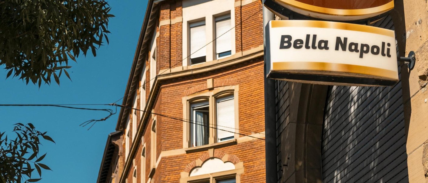 Ein Schild mit der Aufschrift 'Bella Napoli' hängt an einem Backsteingebäude. Der Himmel ist klar und blau, ein Baum ist im Vordergrund sichtbar., © SMG, Sarah Schmid Ein Schild mit der Aufschrift 'Bella Napoli' hängt an einem Backsteingebäude. Der Himmel ist klar und blau, ein Baum ist im Vordergrund sichtbar., © SMG, Sarah Schmid
