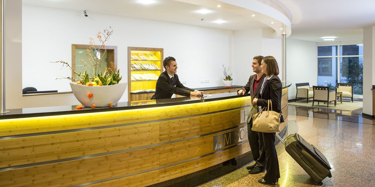 Zwei Gäste stehen an der Rezeption des Best Western Plus Hotel am Schlossberg und sprechen mit einem Mitarbeiter. Im Hintergrund ist eine Sitzlounge sichtbar., © Best Western Plus Hotel am Schlossberg
