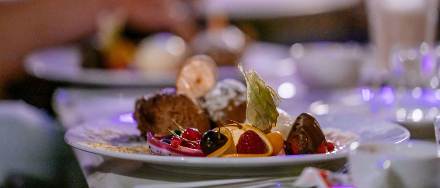 Ein kunstvoll angerichtetes Dessert mit Früchten, Schokolade und Dekoration auf einem Teller. Im Hintergrund unscharfe Lichter., © Thomas Niedermüller