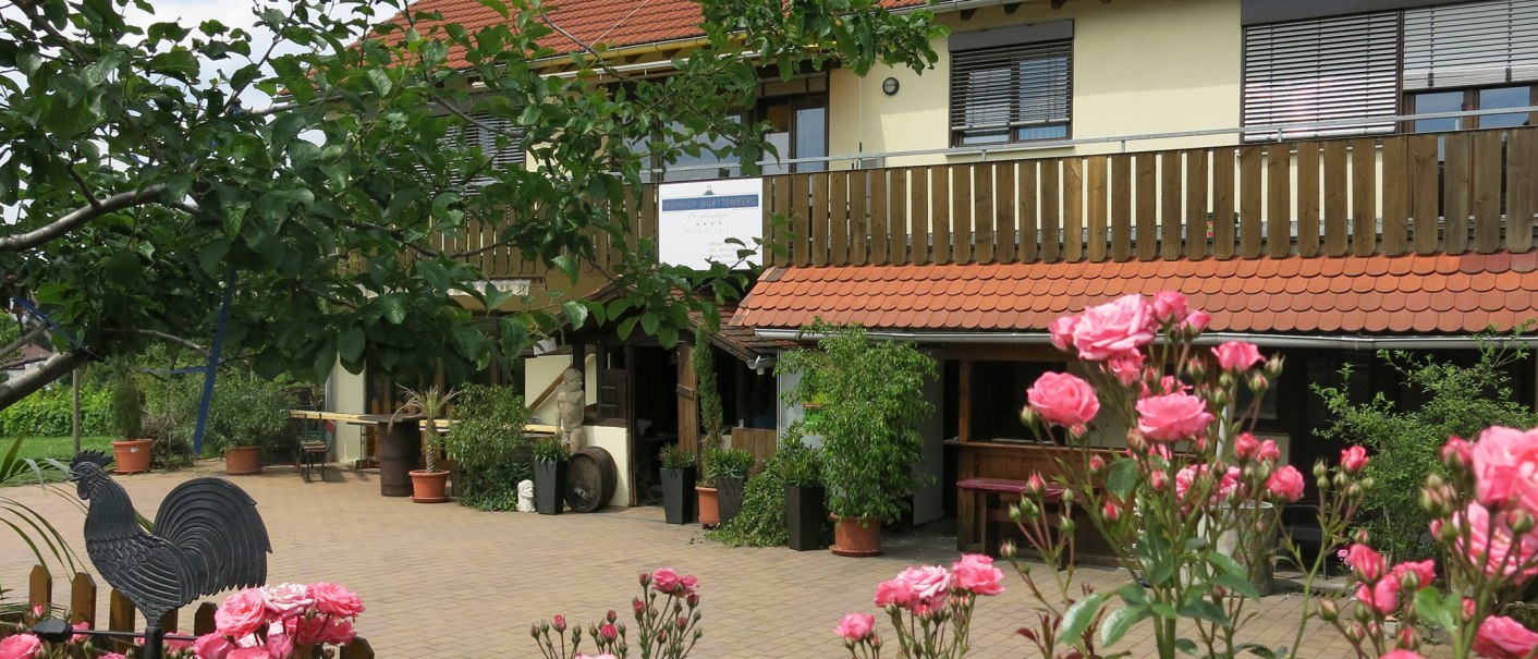 Ein Weinhof mit Holzbalkon, roten Dachziegeln und blühenden Rosen im Vordergrund. Ein Schild mit der Aufschrift 'Weinhof Württemberg Helmut Zaiß' ist sichtbar., © Helmut Zaiß Weingut & Weingastronomie Ein Weinhof mit Holzbalkon, roten Dachziegeln und blühenden Rosen im Vordergrund. Ein Schild mit der Aufschrift 'Weinhof Württemberg Helmut Zaiß' ist sichtbar., © Helmut Zaiß Weingut & Weingastronomie