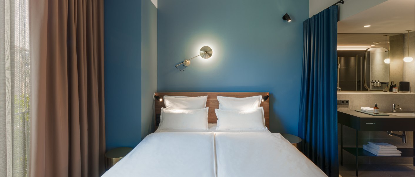 Modernes Hotelzimmer mit Doppelbett, blauer Wand, stilvoller Beleuchtung und angrenzendem Badezimmer. Vorh&auml;nge in Braun und Blau trennen die Bereiche., &copy; EmiLu Hotel GmbH