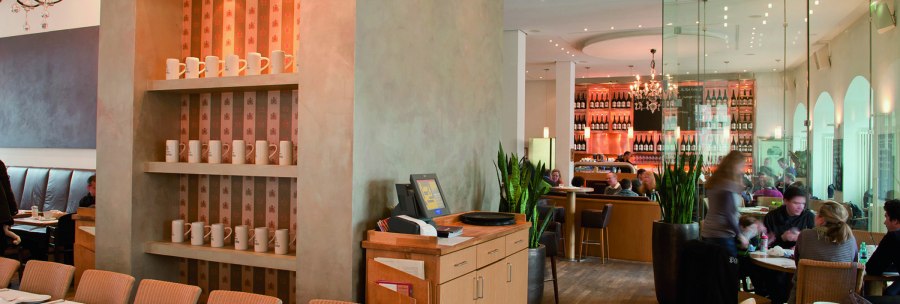 Stilvolles Restaurant mit Holzmöbeln, Regalen mit Tassen und einer Bar im Hintergrund. Menschen sitzen an Tischen, moderne Beleuchtung und Pflanzen dekorieren den Raum., © aspectus photographie, Stuttgart