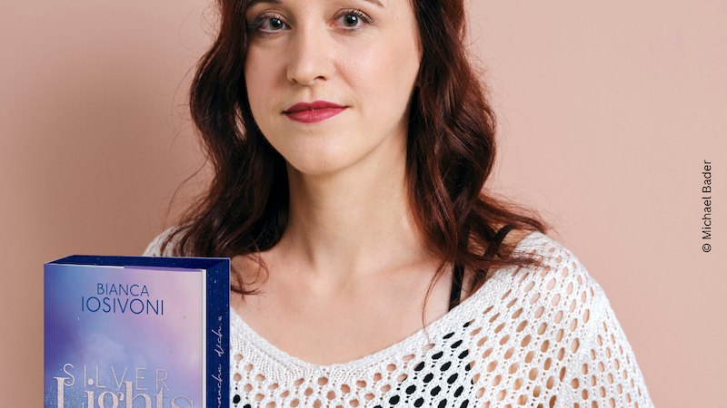 Eine Frau mit langen, dunklen Haaren steht vor einem rosa Hintergrund. Neben ihr ist das Buch 'Silver Lights - The more I love you' von Bianca Iosivoni zu sehen., &copy; Thalia Deutschland GmbH & Co. KG
