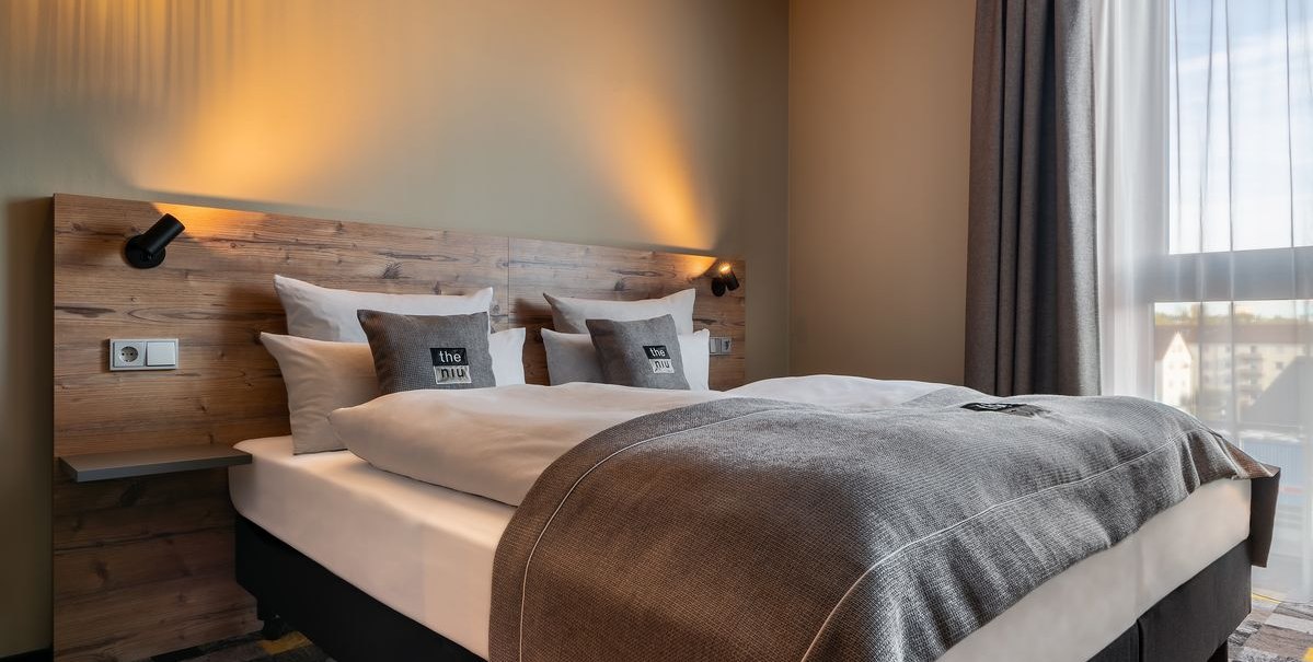 Modernes Hotelzimmer mit Doppelbett, Holzkopfteil, grauen Kissen und Vorhängen. Warmes Licht beleuchtet den Raum., © Novum Hospitality