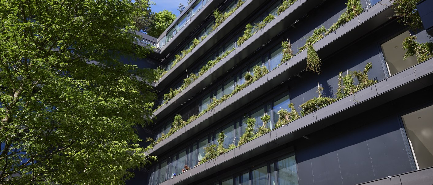 Modernes Gebäude mit begrünten Balkonen, Pflanzen wachsen entlang der Fassade. Ein Baum im Vordergrund, blauer Himmel im Hintergrund., © Ferdinand Piëch Holding GmbH