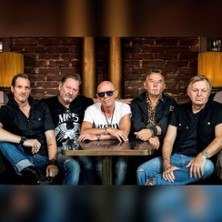 Fünf Männer sitzen lässig an einem Tisch vor einer Backsteinwand. Sie tragen überwiegend schwarze Kleidung und wirken entspannt., © links im Bild