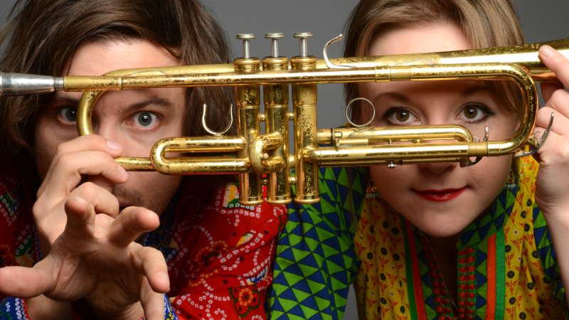 Zwei Personen in bunten Hemden blicken durch eine Trompete. Sie halten das Instrument vor ihre Gesichter und schauen neugierig hindurch., © Matria Zwei Personen in bunten Hemden blicken durch eine Trompete. Sie halten das Instrument vor ihre Gesichter und schauen neugierig hindurch., © Matria