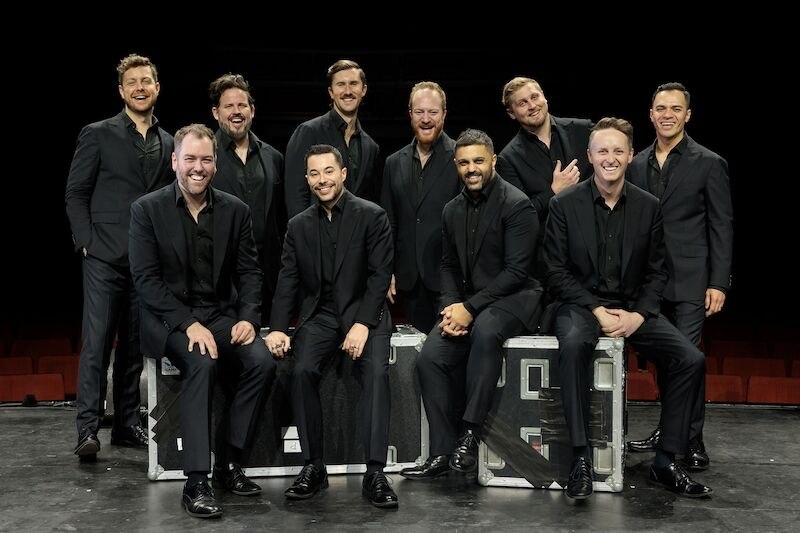Ten men in black suits pose smiling on a stage. Some are sitting on boxes, others are standing behind them. They appear cheerful and relaxed., &copy; Theaterhaus Stuttgart e.V.