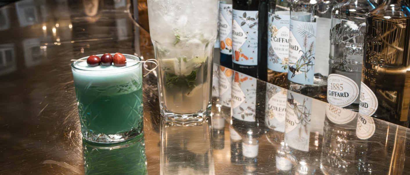 Two cocktails on a shiny bar. On the left a blue drink with berries, on the right a mojito. In the background are bottles of liqueurs., © SMG Stuttgart Marketing GmbH - Sarah Schmid Two cocktails on a shiny bar. On the left a blue drink with berries, on the right a mojito. In the background are bottles of liqueurs., © SMG Stuttgart Marketing GmbH - Sarah Schmid