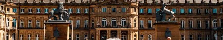 Das Neue Schloss Stuttgart bei sonnigem Wetter. Menschen gehen und ein Fahrradfahrer f&auml;hrt im Vordergrund. Zwei Statuen flankieren den Eingang., &copy; Stuttgart-Marketing GmbH, Sarah Schmid