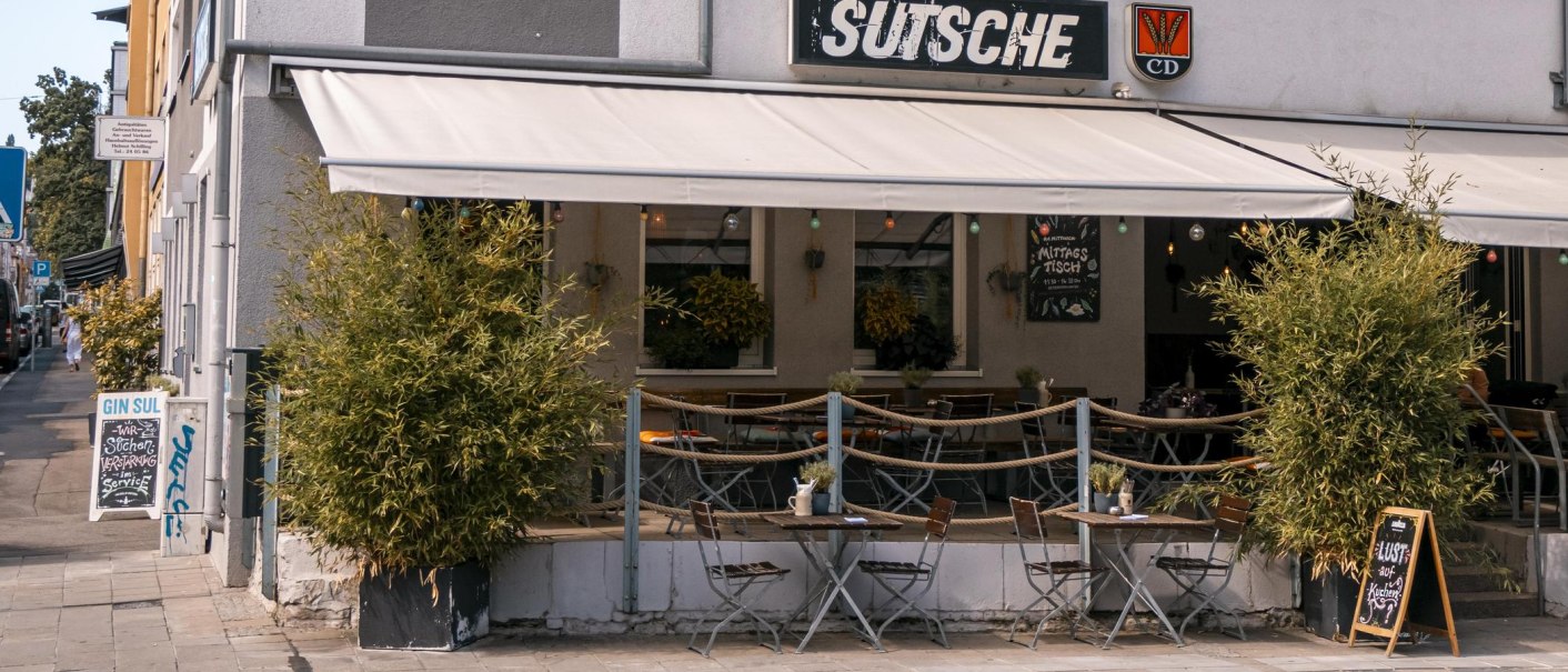 Café 'Sutsche' mit Außenbereich, Tischen und Stühlen auf dem Gehweg, umgeben von Pflanzen., © SMG, Sarah Schmid