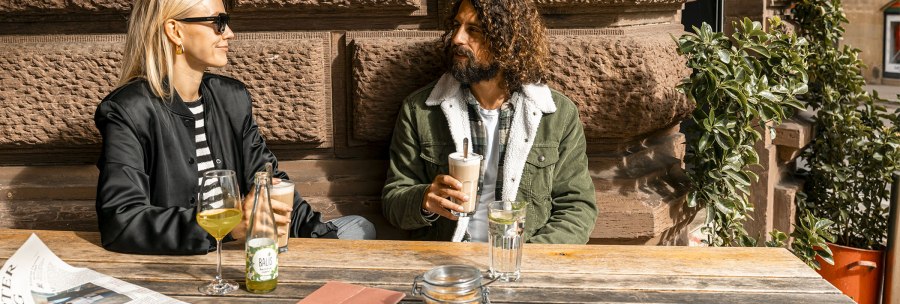 Zwei Personen sitzen an einem Holztisch vor einer Steinmauer, trinken Getr&auml;nke und unterhalten sich. Eine Zeitung liegt auf dem Tisch., &copy; SMG, Sarah Schmid
