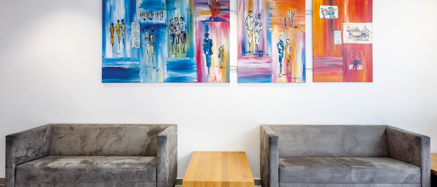 Zwei graue Sofas stehen gegen&uuml;ber, getrennt durch einen Holztisch. An der wei&szlig;en Wand h&auml;ngen bunte, abstrakte Gem&auml;lde., &copy; Seminaris Hotel Bad Boll