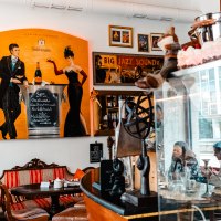 Ein gemütliches Café mit bunten Postern, darunter ein großes Bild mit einem Paar und Champagner. Menschen sitzen an Tischen, umgeben von Dekorationen., © SMG_Sarah Schmid