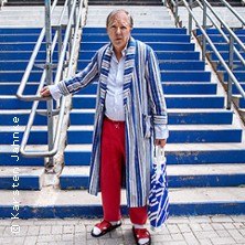 Person in gestreiftem Bademantel und roter Hose steht vor blauen Treppen, hält eine Tasche und trägt Sandalen., © links im Bild Person in gestreiftem Bademantel und roter Hose steht vor blauen Treppen, hält eine Tasche und trägt Sandalen., © links im Bild