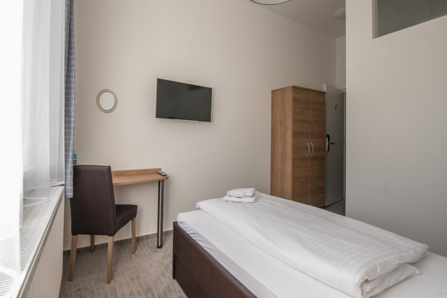 Ein schlichtes Einzelzimmer mit Bett, Schreibtisch, Stuhl, Wandfernseher und Kleiderschrank. Helle Wände und Fenster mit Vorhang., © TOMAS Ein schlichtes Einzelzimmer mit Bett, Schreibtisch, Stuhl, Wandfernseher und Kleiderschrank. Helle Wände und Fenster mit Vorhang., © TOMAS