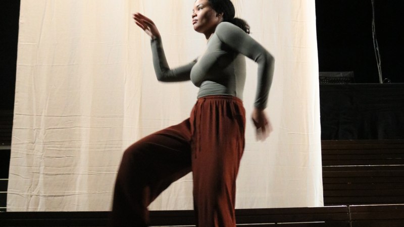 A person is dancing on a stage in front of a large, light-colored curtain. She is wearing a gray top and brown pants., &copy; Amina Ousman