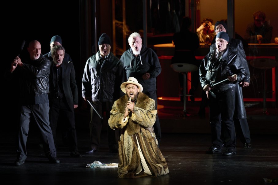 Ein Mann in Pelzmantel und Hut kniet auf der Bühne, umgeben von Männern in dunkler Kleidung. Dramatische Opernszene mit intensiver Beleuchtung., © Martin Sigmund