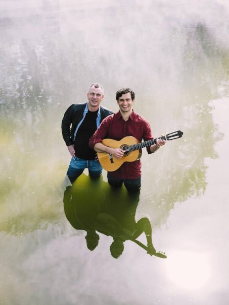 Two men are standing up to their knees in a pond. One is carrying a guitar and smiling. Their reflections are visible in the water., &copy; Rosenau Kultur e.V.