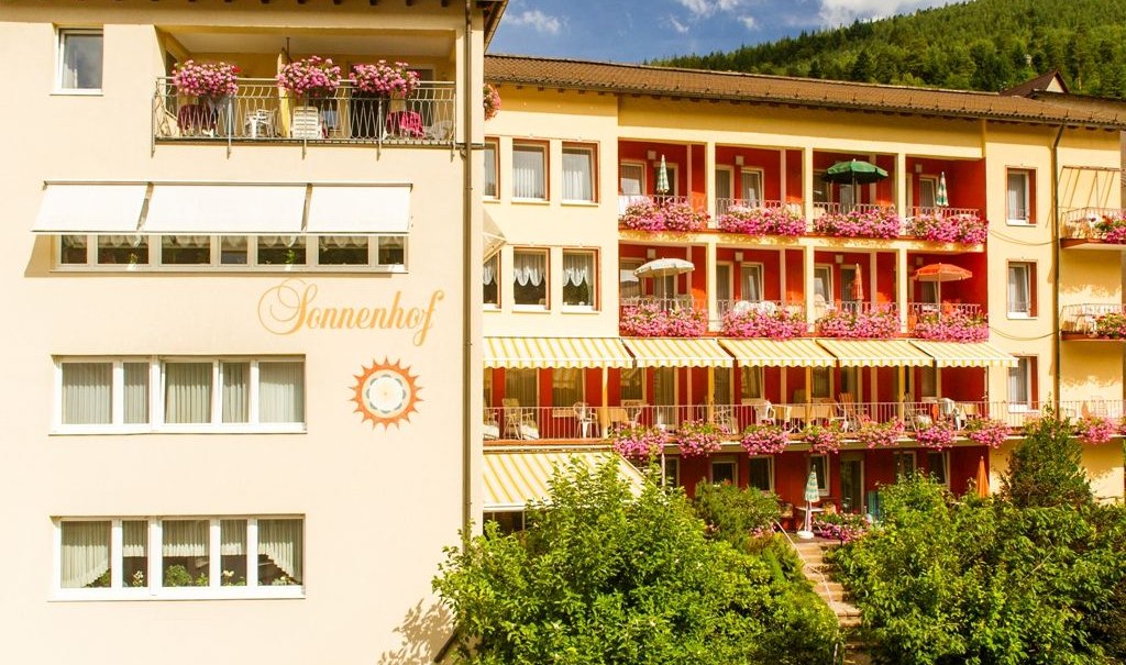 Das Hotel Sonnenhof zeigt eine Fassade mit blühenden Balkonen und Sonnenschirmen. Der Schriftzug 'Sonnenhof' ist sichtbar. Im Hintergrund sind Bäume zu sehen., © Hotel Sonnenhof