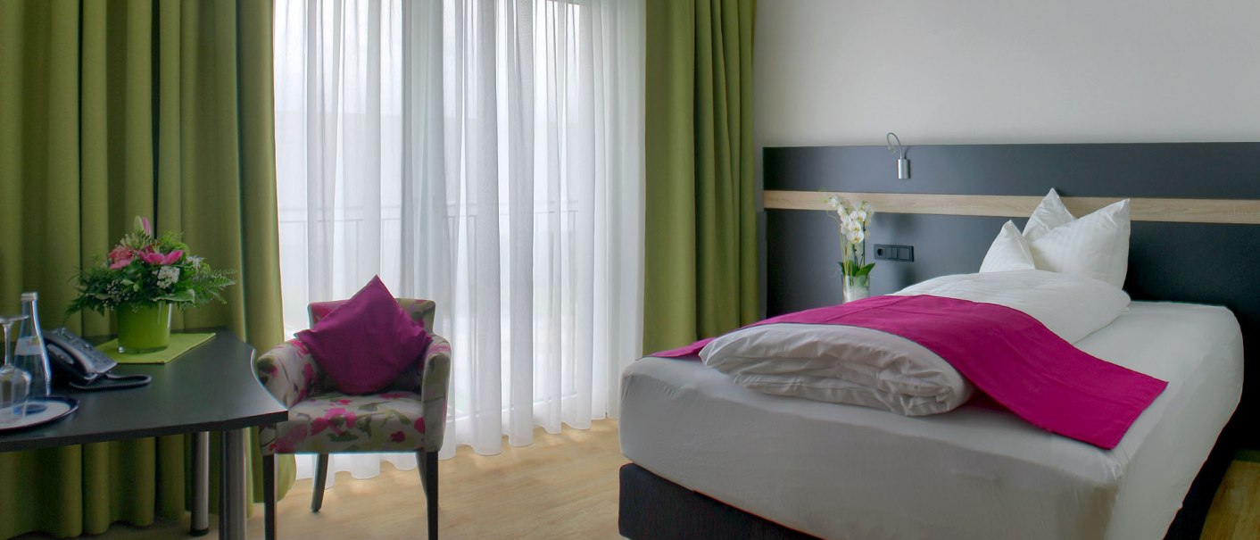 A modern single room with a bed, a table with flowers, a chair and green curtains in front of a window., © TOMAS