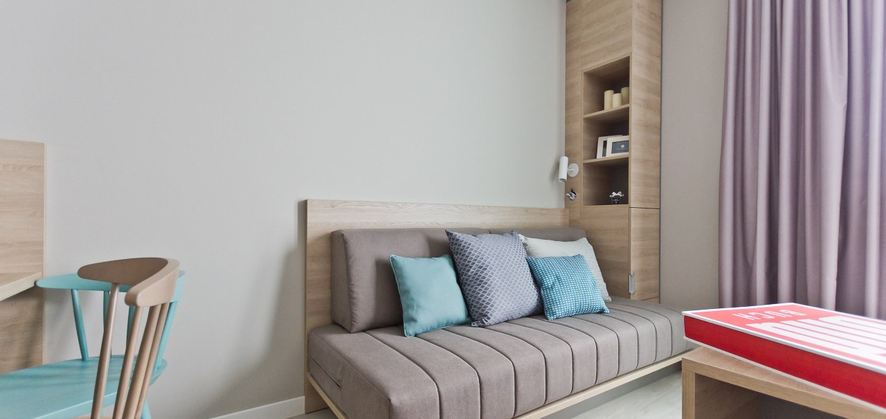 Modernes Apartment mit grauem Sofa, bunten Kissen, Holzm&ouml;beln und lila Vorh&auml;ngen. Ein Tisch mit rotem Buch ist im Vordergrund sichtbar., &copy; Arthotel Ana / elaya