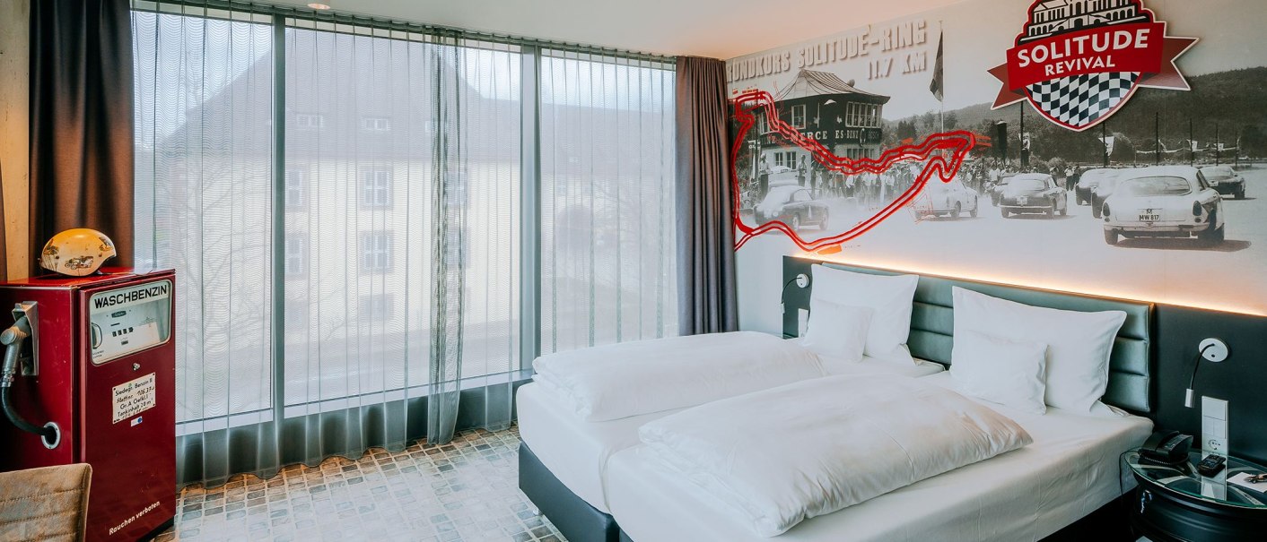 Hotelzimmer im V8 HOTEL mit Rennstrecken-Dekor, Doppelbett, Retro-Tankstelle und großem Fenster. An der Wand historische Rennfotos und Solitude Revival Logo., © V8 HOTEL