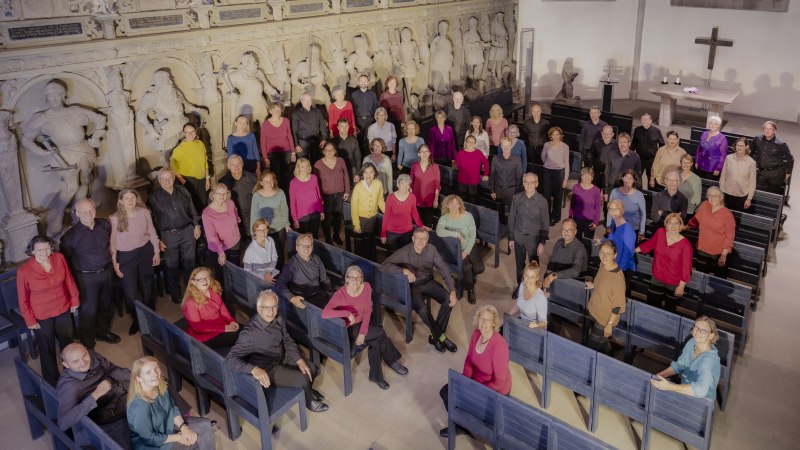 Eine große Gruppe von Menschen steht in einer Kirche vor einer Wand mit historischen Reliefs. Sie tragen bunte Kleidung und lächeln in die Kamera., © Stuttgarter Kantorei Eine große Gruppe von Menschen steht in einer Kirche vor einer Wand mit historischen Reliefs. Sie tragen bunte Kleidung und lächeln in die Kamera., © Stuttgarter Kantorei