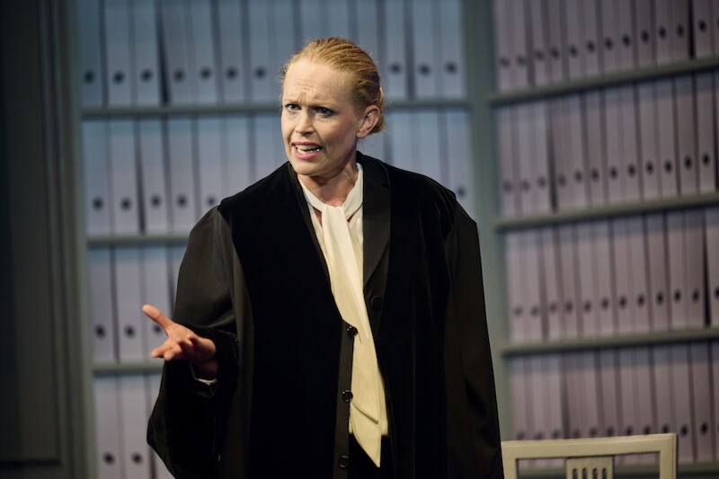 A woman in a black lawyer's coat gestures in front of a shelf full of files. She looks committed and focused., &copy; Theaterhaus Stuttgart e.V.