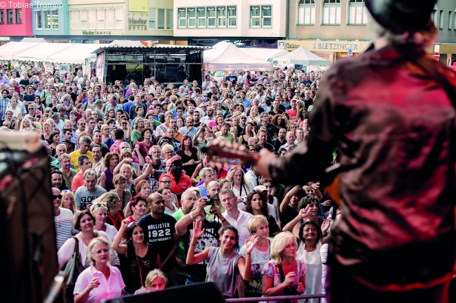 Ein Musiker spielt auf einer Bühne vor einer großen, begeisterten Menschenmenge bei einem Festival in einer Stadt., © Tobias Trumpp Ein Musiker spielt auf einer Bühne vor einer großen, begeisterten Menschenmenge bei einem Festival in einer Stadt., © Tobias Trumpp