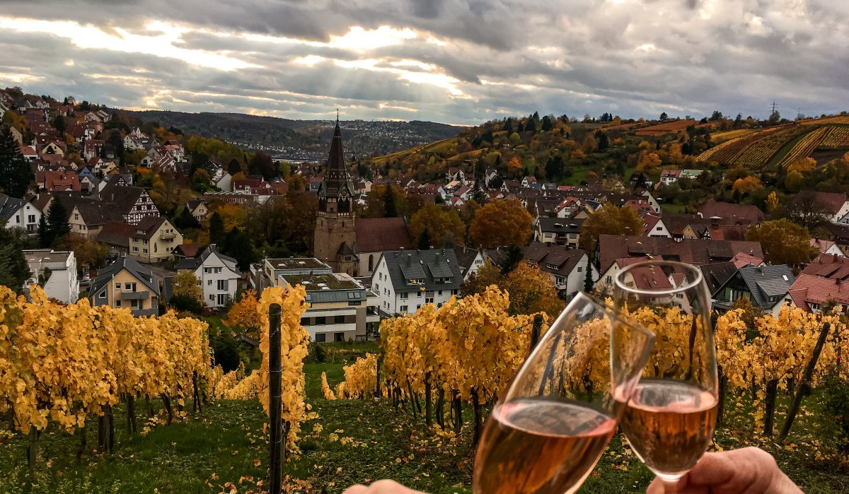Two people toast with wine glasses in an autumnal vineyard. A town with a church and colorful trees can be seen in the background., © Stuttgart-Marketing GmbH