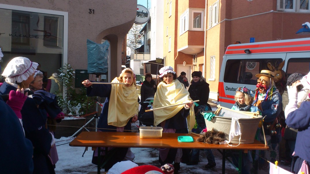 Straßenfest mit verkleideten Menschen, die traditionelle Kostüme tragen. Ein Waschbrett und ein Eimer stehen im Mittelpunkt der Szene., © Narrenzunft Feuerbach e. V. Straßenfest mit verkleideten Menschen, die traditionelle Kostüme tragen. Ein Waschbrett und ein Eimer stehen im Mittelpunkt der Szene., © Narrenzunft Feuerbach e. V.
