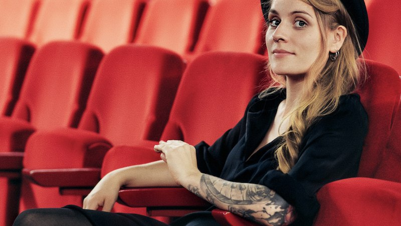 Eine Frau mit Hut und tätowiertem Arm sitzt entspannt in einem leeren Kino auf roten Sitzen., © Renitenztheater Stuttgart e.V. Eine Frau mit Hut und tätowiertem Arm sitzt entspannt in einem leeren Kino auf roten Sitzen., © Renitenztheater Stuttgart e.V.